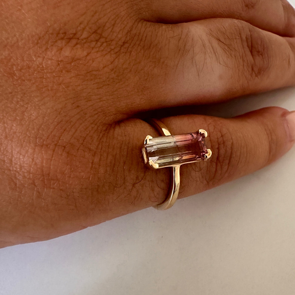 A gold ring with a rectangular pinkish gemstone worn on a person's finger, shown against a plain light background.